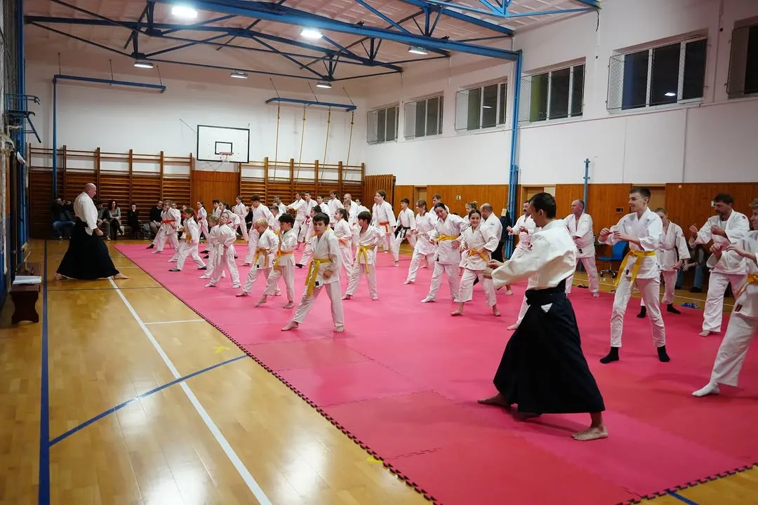 Deti v kimonách s farebnými pásmi cvičia na tatami počas skúšok, na lavičkách sedia rodičia